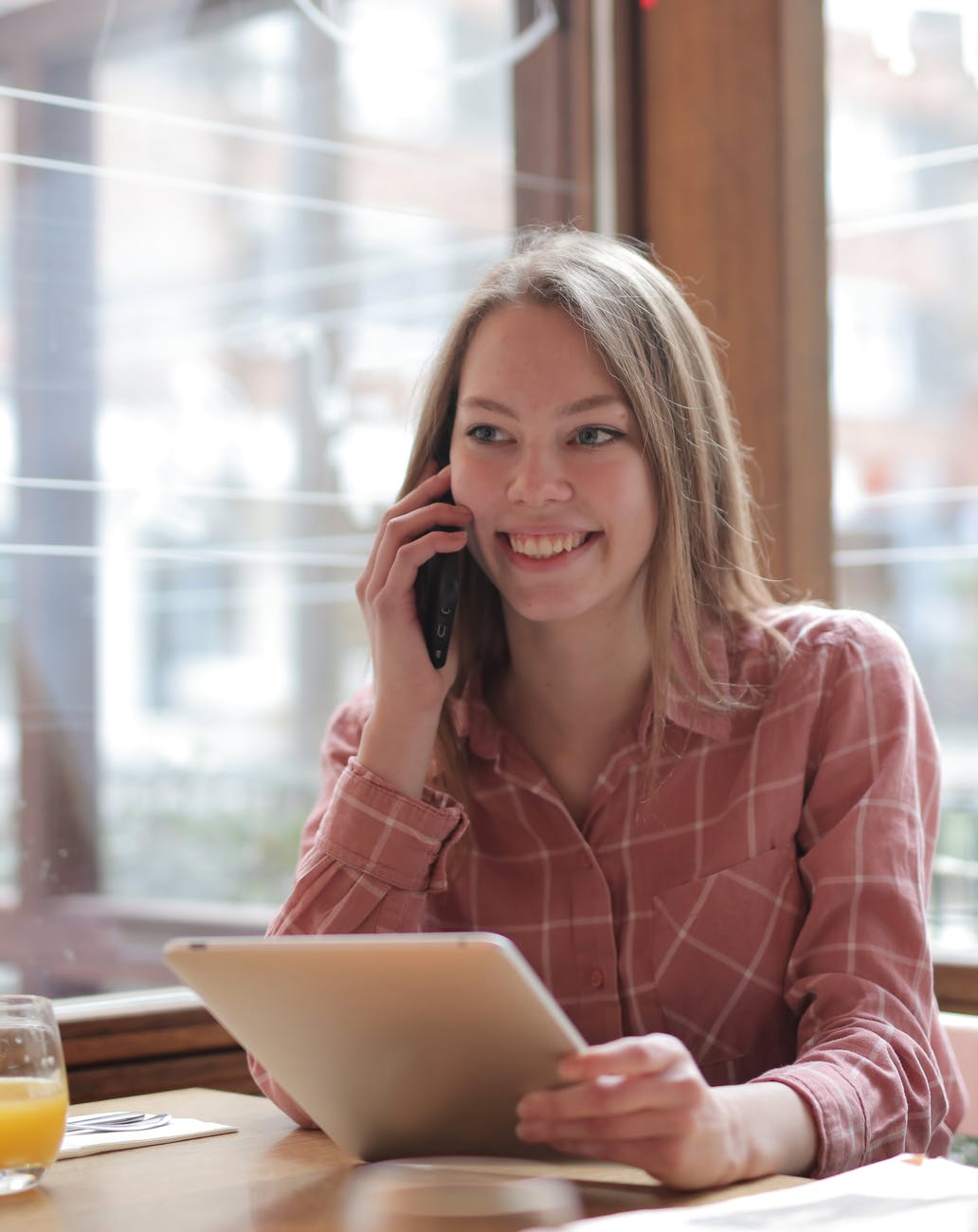 En la imagen, una mujer sonriente situada en una cafetería, porta en una mano una tablet, y con la otra está realizando una llamada de teléfono a FUNTESO, Fundación Tecnología Social, 