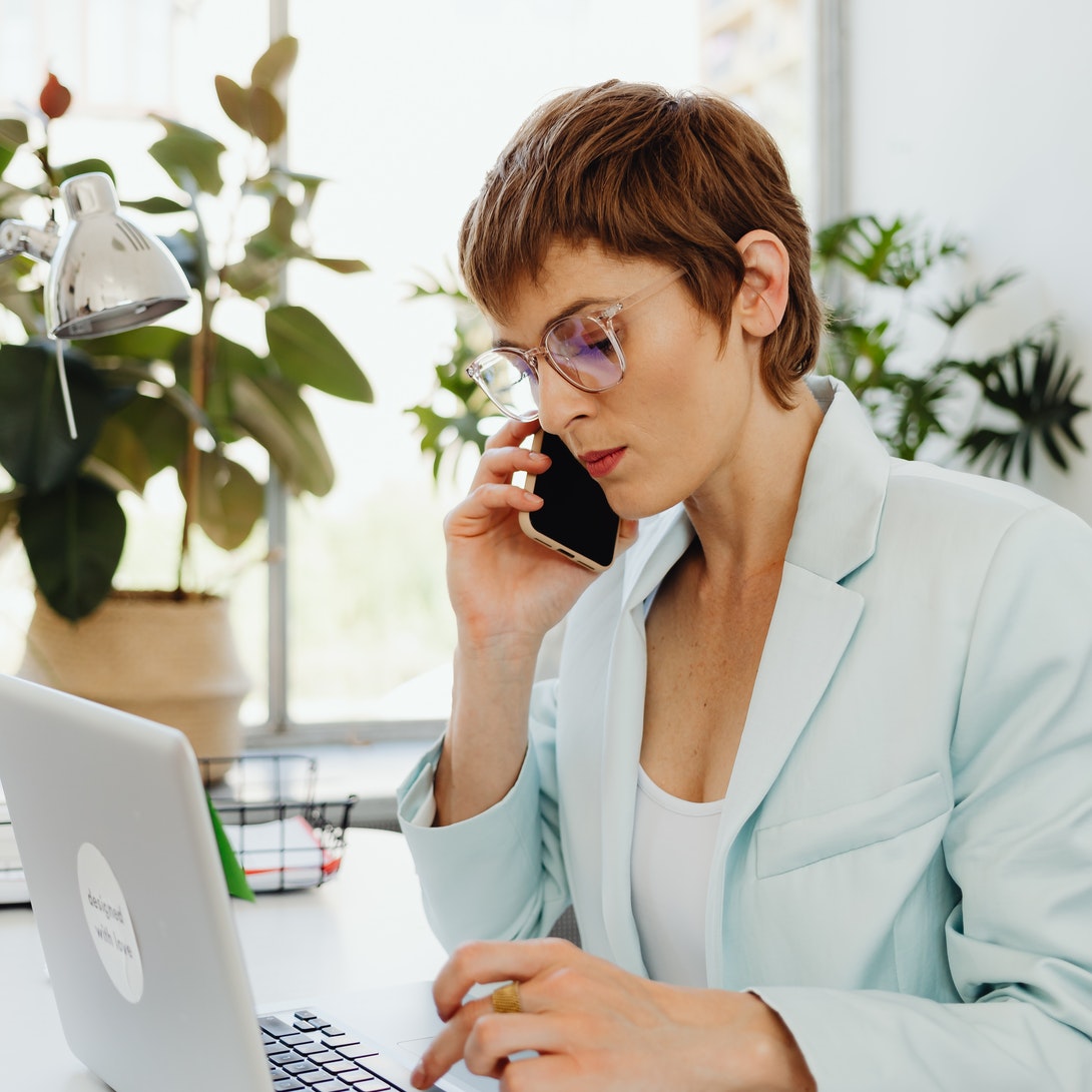 En la imagen aparece una mujer llamando a través de un teléfono móvil. La mujer se encuentra sentada, frente a un ordenador portátil con la pantalla abierta.